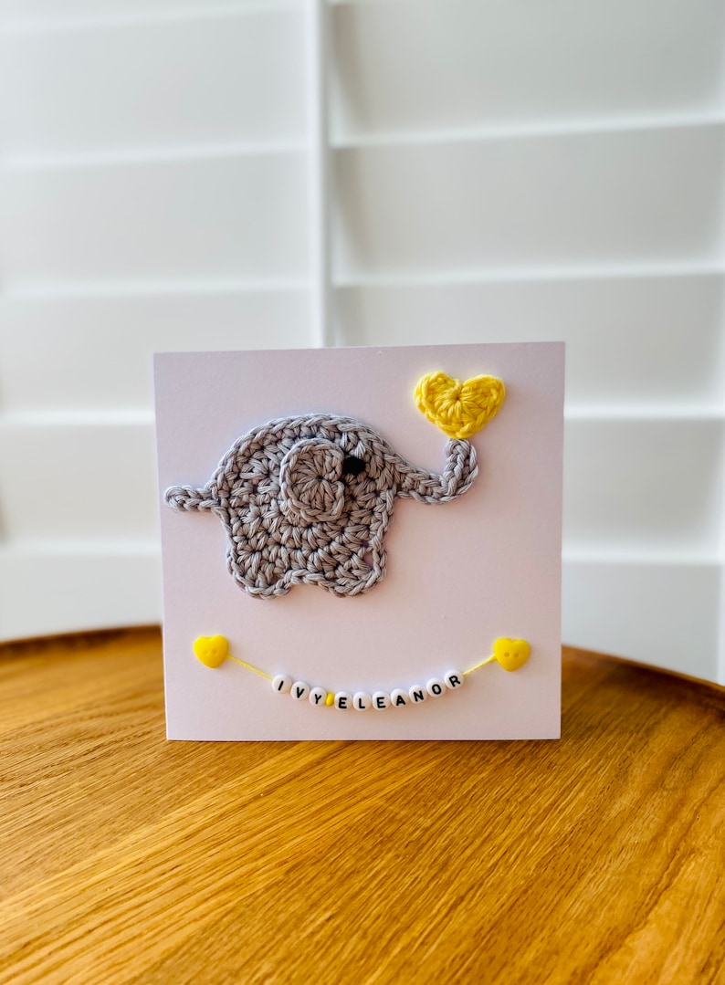 Puede incluir: Un elefante de crochet gris que sostiene un coraz&oacute;n de crochet amarillo. El elefante est&aacute; en una tarjeta blanca con una cadena de cuentas amarillas que deletrean "IVY ELEANOR".