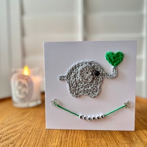 Puede incluir: Una tarjeta blanca con un elefante de crochet gris que sostiene un coraz&oacute;n verde. El elefante est&aacute; en una tarjeta blanca con una cuerda verde con estrellas plateadas y el nombre "GEORGE" escrito en cuentas blancas.