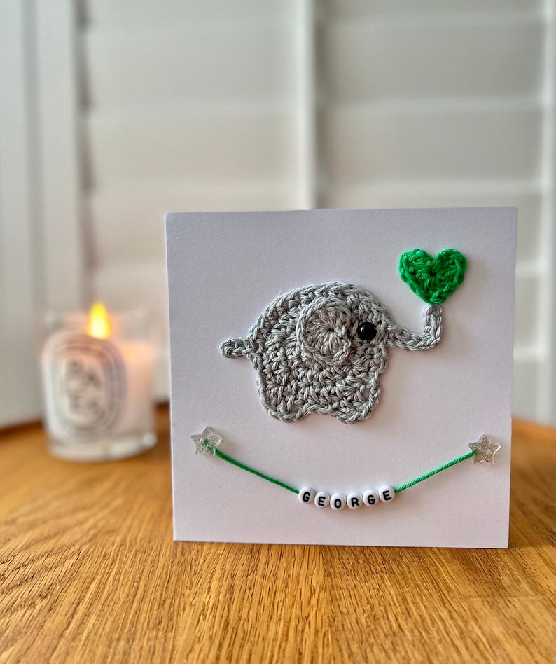 Puede incluir: Una tarjeta blanca con un elefante de crochet gris que sostiene un coraz&oacute;n verde. El elefante est&aacute; en una tarjeta blanca con una cuerda verde con estrellas plateadas y el nombre "GEORGE" escrito en cuentas blancas.