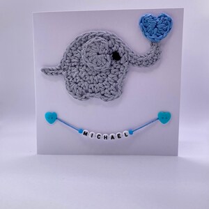 Puede incluir: Un elefante de crochet gris sosteniendo un coraz&oacute;n de crochet azul. Una cuerda azul con cuentas de coraz&oacute;n azul deletrea el nombre "MICHAEL" en una tarjeta blanca.