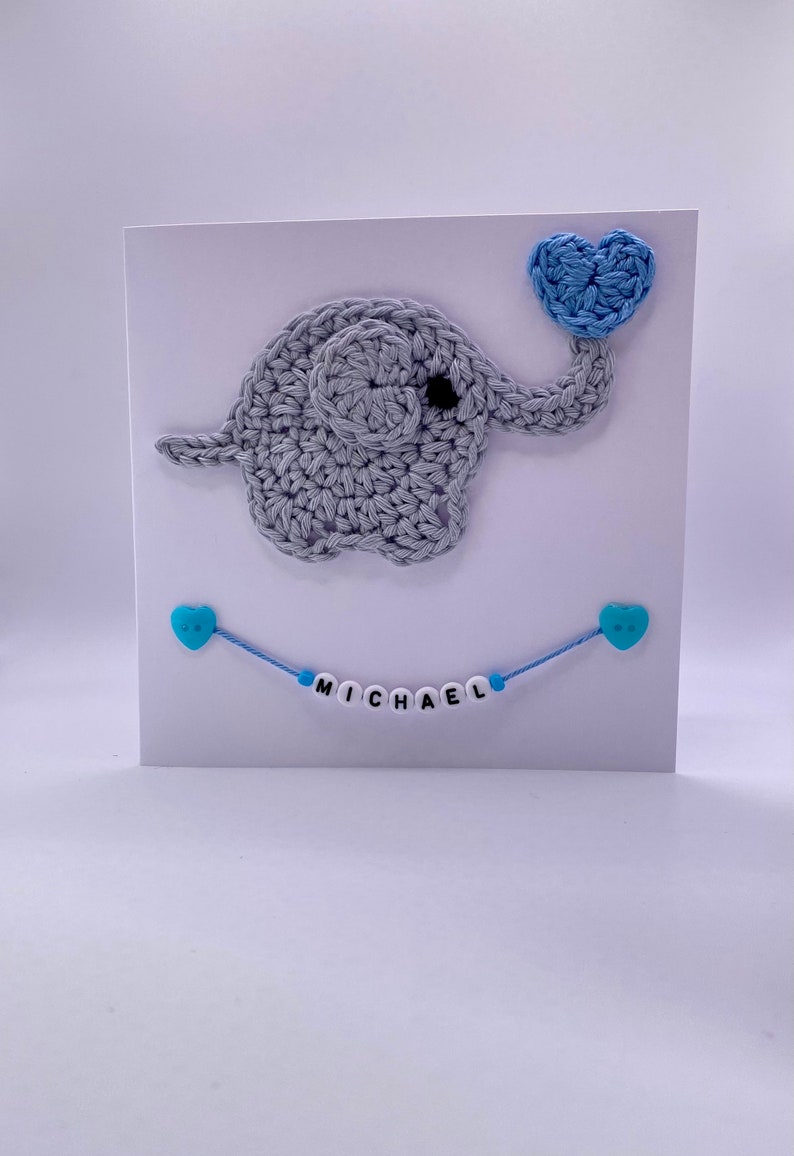 Puede incluir: Un elefante de crochet gris sosteniendo un coraz&oacute;n de crochet azul. Una cuerda azul con cuentas de coraz&oacute;n azul deletrea el nombre "MICHAEL" en una tarjeta blanca.