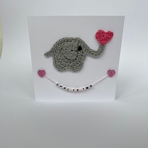 Puede incluir: Un elefante de crochet gris sosteniendo un coraz&oacute;n de crochet rosa. El elefante est&aacute; en una tarjeta blanca con corazones rosas y una cuerda rosa y blanca con cuentas blancas que deletrean "Opal is Two".