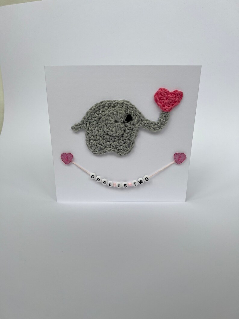 Puede incluir: Un elefante de crochet gris sosteniendo un coraz&oacute;n de crochet rosa. El elefante est&aacute; en una tarjeta blanca con corazones rosas y una cuerda rosa y blanca con cuentas blancas que deletrean "Opal is Two".