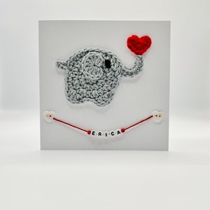 Puede incluir: Un elefante de crochet gris que sostiene un coraz&oacute;n rojo con el nombre "ERICA" escrito en cuentas blancas en una cuerda roja.