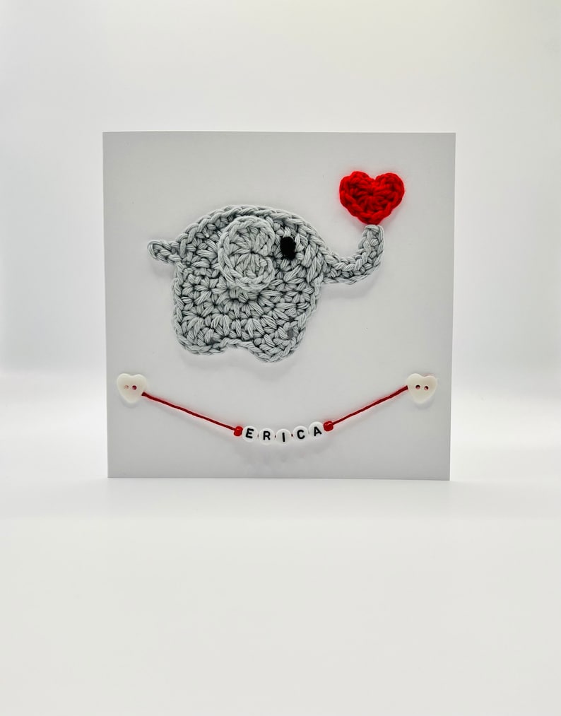 Puede incluir: Un elefante de crochet gris que sostiene un coraz&oacute;n rojo con el nombre "ERICA" escrito en cuentas blancas en una cuerda roja.
