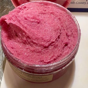 May include: A clear jar containing a bright pink body scrub. The scrub has a textured, grainy appearance, indicating exfoliating properties. The container allows the product's colour to be seen. The label is partially visible, with text that is difficult to read.
