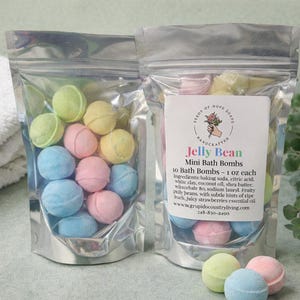 May include: Two clear bags filled with colorful, round bath bombs. The bath bombs are in shades of blue, pink, yellow, and green. The bags have a label that reads "Jelly Bean Mini Bath Bombs". The label also lists the ingredients and the website address.