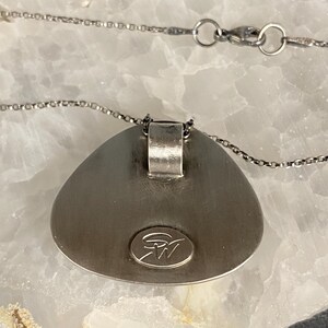 May include: A silver pendant necklace with a teardrop-shaped pendant. The pendant has a smooth, rounded surface and a small, circular logo on the back. The necklace is made of a delicate silver chain.