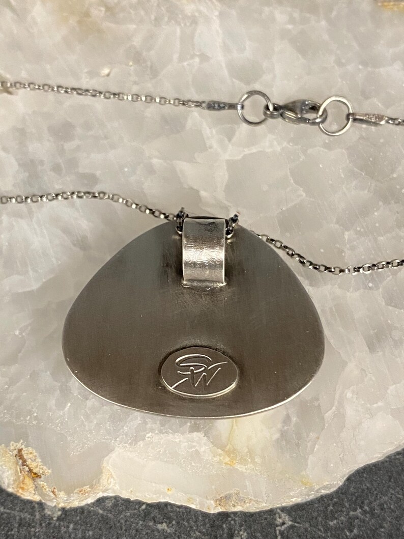 May include: A silver pendant necklace with a teardrop-shaped pendant. The pendant has a smooth, rounded surface and a small, circular logo on the back. The necklace is made of a delicate silver chain.