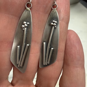 May include: A pair of silver-toned earrings with a botanical design. Each earring features a long, teardrop-shaped plate with a textured surface. The design includes a cluster of small, round beads and vertical bar accents.