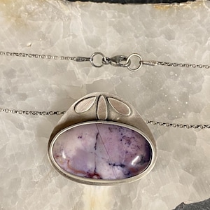 May include: A silver pendant necklace with a purple and white agate stone set in a silver oval frame. The pendant has a delicate silver design with four small leaves at the top.