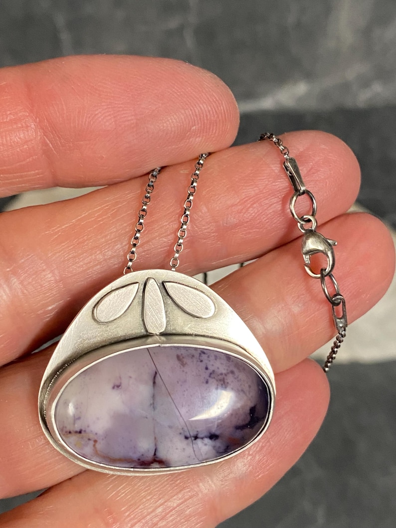 May include: A silver pendant necklace with a purple and white agate stone set in a silver bezel. The pendant has a delicate design with four small silver leaves around the stone.