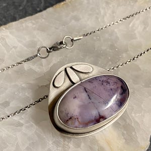 May include: A silver pendant necklace with a purple and white agate stone set in a silver frame. The pendant has a geometric design with three silver leaves on the top.