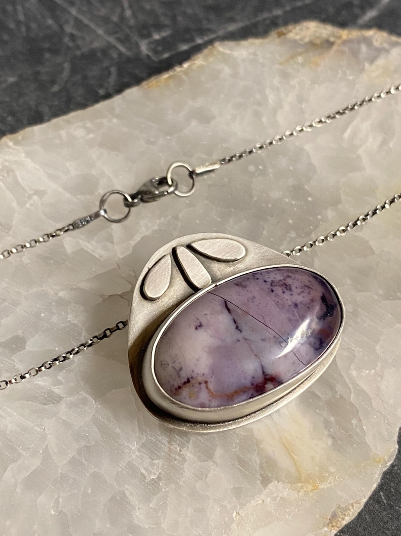 May include: A silver pendant necklace with a purple and white agate stone set in a silver frame. The pendant has a geometric design with three silver leaves on the top.