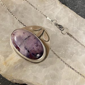 May include: A silver necklace with a purple and white stone pendant. The pendant is set in a silver frame with a floral design.