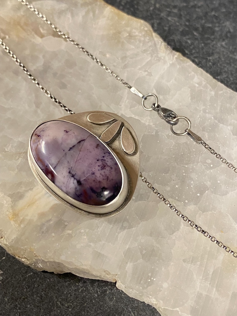 May include: A silver necklace with a purple and white stone pendant. The pendant is set in a silver frame with a floral design.