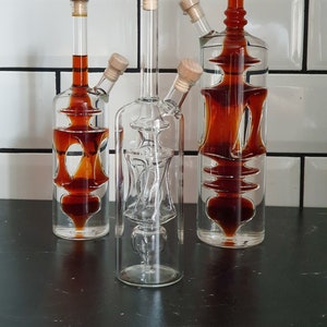 K&ouml;nnte beinhalten: Drei Glasflaschen mit Korkstopfen und einem einzigartigen Design. Die Flaschen sind mit einer dunkelbraunen Fl&uuml;ssigkeit gef&uuml;llt. Die Flaschen stehen auf einer schwarzen Oberfl&auml;che.
