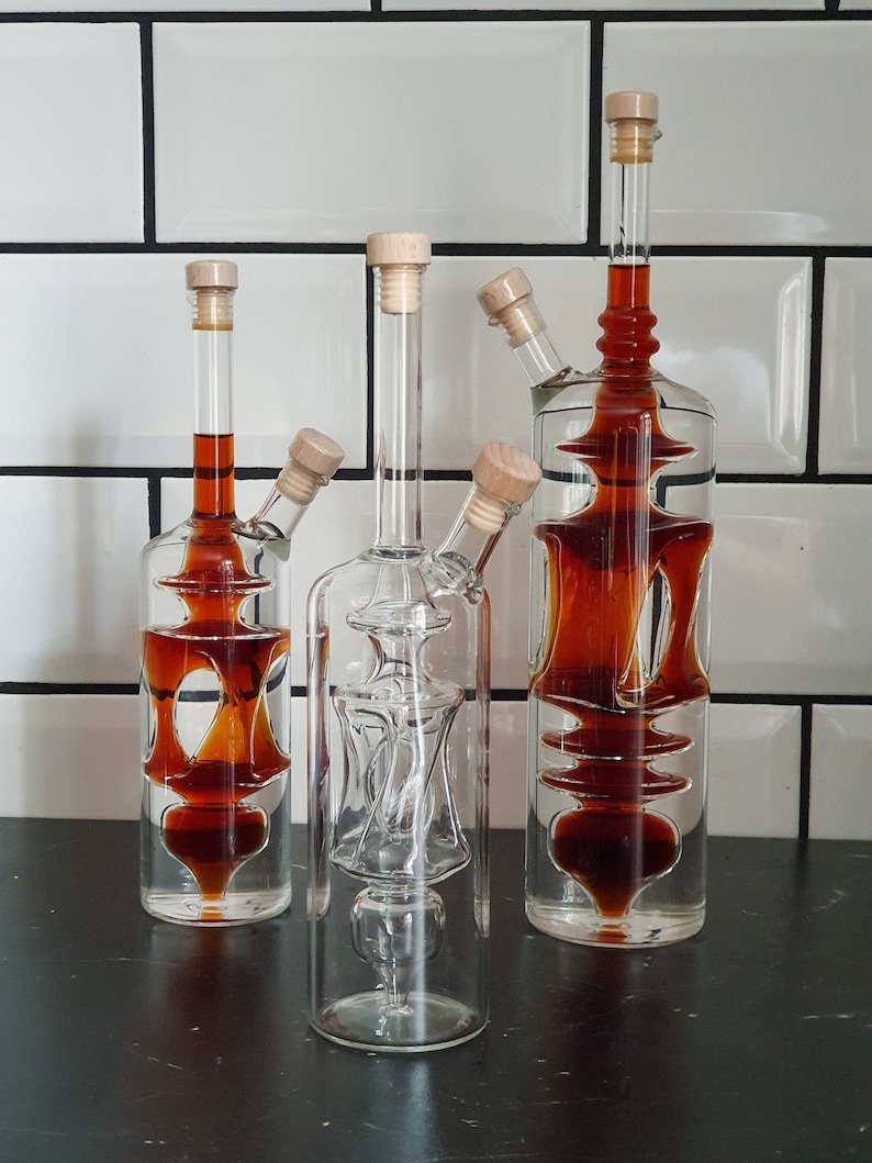 K&ouml;nnte beinhalten: Drei Glasflaschen mit Korkstopfen und einem einzigartigen Design. Die Flaschen sind mit einer dunkelbraunen Fl&uuml;ssigkeit gef&uuml;llt. Die Flaschen stehen auf einer schwarzen Oberfl&auml;che.