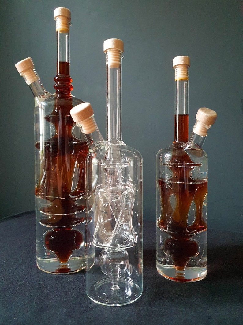 K&ouml;nnte beinhalten: Drei Glasflaschen mit einzigartigen, komplizierten Designs. Die Flaschen sind aus klarem Glas mit einer braunen Fl&uuml;ssigkeit im Inneren. Die Flaschen haben Holzstopfen.