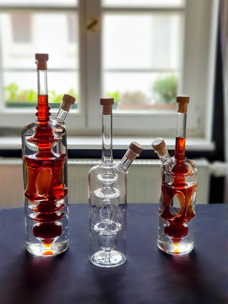 K&ouml;nnte beinhalten: Drei Glasflaschen mit einem einzigartigen Design, jede mit einem Korkstopfen und gef&uuml;llt mit einer dunkelroten Fl&uuml;ssigkeit. Die Flaschen stehen auf einer schwarzen Oberfl&auml;che.