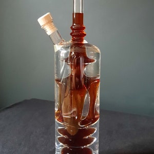 K&ouml;nnte beinhalten: Eine klare Glasflasche mit zwei Ausg&uuml;ssen und einem Holzstopfen. Die Flasche ist mit einer dunkelbraunen Fl&uuml;ssigkeit gef&uuml;llt und hat ein einzigartiges, geschichtetes Design.