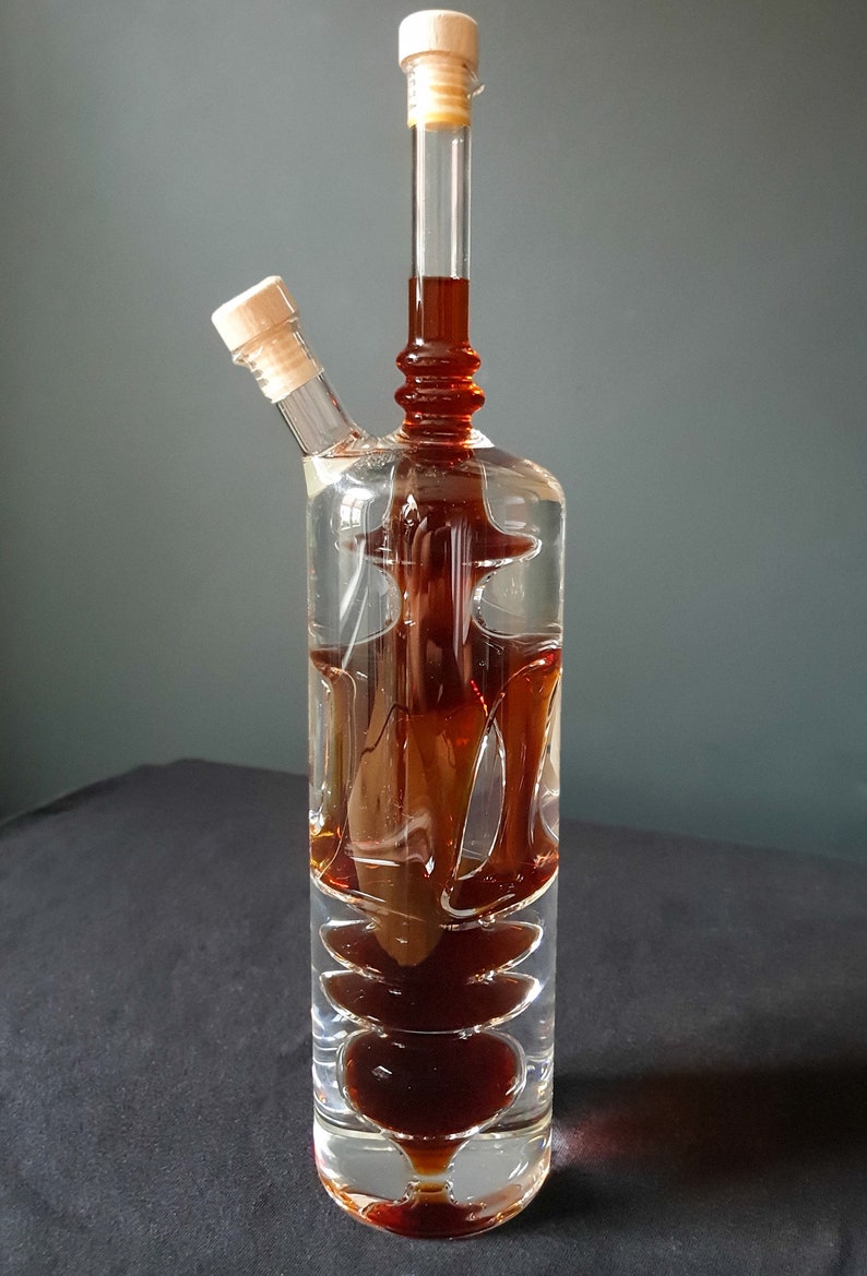 K&ouml;nnte beinhalten: Eine klare Glasflasche mit zwei Ausg&uuml;ssen und einem Holzstopfen. Die Flasche ist mit einer dunkelbraunen Fl&uuml;ssigkeit gef&uuml;llt und hat ein einzigartiges, geschichtetes Design.
