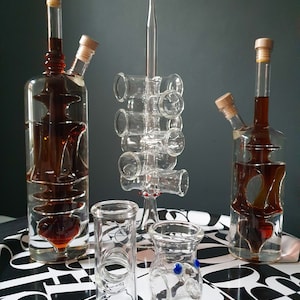 K&ouml;nnte beinhalten: Zwei klare Glasflaschen mit brauner Fl&uuml;ssigkeit und Korkstopfen, ein Glas-Schnapsglashalter mit sechs Schnapsgl&auml;sern und zwei klare Glas-Schnapsgl&auml;ser mit blauen und roten Akzenten auf einer schwarz-wei&szlig;en gemusterten Oberfl&auml;che.