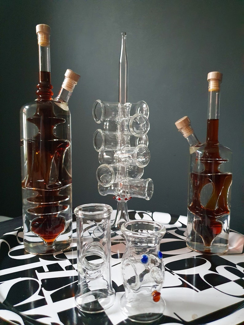 K&ouml;nnte beinhalten: Zwei klare Glasflaschen mit brauner Fl&uuml;ssigkeit und Korkstopfen, ein Glas-Schnapsglashalter mit sechs Schnapsgl&auml;sern und zwei klare Glas-Schnapsgl&auml;ser mit blauen und roten Akzenten auf einer schwarz-wei&szlig;en gemusterten Oberfl&auml;che.