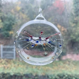 Op de afbeelding: Een helder, bolvormig glazen ornament met een interne structuur van fijne, uitstralende glazen stekels. De stekels zijn voorzien van kleine, levendige stippen in rood, blauw en goud, die in de holle glazen bol zijn opgehangen.