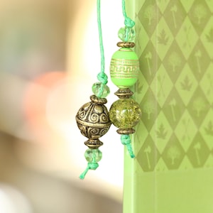 Puede incluir: Primer plano de un marcapáginas con cuentas verdes y doradas. El marcapáginas presenta cuentas decorativas en tonos verdes y dorados, ensartadas en un cordón verde. El fondo es un libro verde claro con un patrón de diamantes.