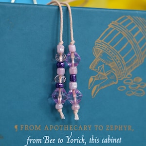 May include: Two beaded bookmarks with pink and purple beads hang in front of a teal book. The beads are strung on a beige cord. The book has gold text and an illustration of a barrel.