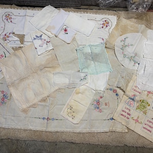May include: A collection of vintage embroidered linens, including tablecloths, napkins, and decorative pieces. The textiles feature floral and geometric patterns in various colors, with some pieces bearing stitched text. The items are arranged on a neutral surface.