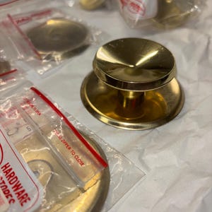 May include: A gold-toned cabinet knob with a round base and a raised, circular top. The knob is packaged in a clear plastic bag with a red label that says "Press Upper to Close".