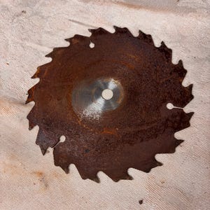 May include: A circular saw blade with a rusted, dark brown finish and sharp, jagged teeth along the outer edge. The center of the metal disc features a smooth, circular hole with a metallic silver finish, showing signs of wear and oxidation.