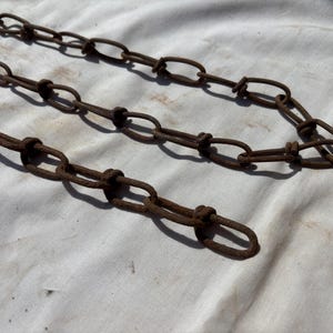 May include: Close-up of three rusty metal chains. The chains are made of oval links connected by smaller, knotted links. The chains are a dark brown color and appear to be old. The background is a white fabric.