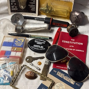 May include: A collection of vintage items including a View-Master reel, a pair of aviator sunglasses, a small red book titled "The Constitution of the United States of America", a key, a seashell, a paint can, a bottle, a small red, white, and blue box, a small metal box, a pen, a pair of pliers, a small metal object, a small metal box, and a small metal object.