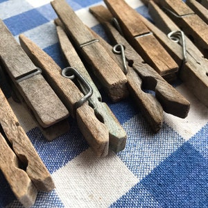 May include: A collection of weathered wooden clothespins with metal springs, arranged on a blue and white checkered fabric. The clothespins are various shades of brown and gray, showing signs of age and use. The metal springs are silver.