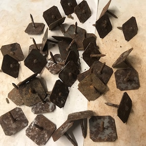 May include: A collection of rusty metal tacks with square heads and pointed tips. The tacks are scattered on a white surface.