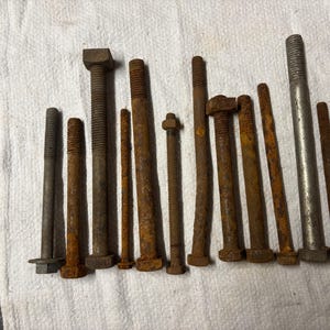 May include: A collection of various sizes of metal bolts, some with hexagonal heads, arranged on a white cloth. The bolts show signs of rust and wear, indicating age and use. The bolts vary in length and diameter.