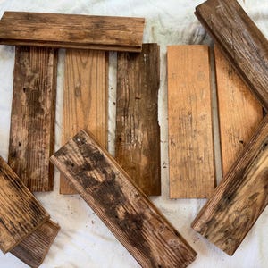 May include: A collection of rectangular wooden planks featuring natural grain patterns, varying shades of brown, and weathered textures. The boards are arranged in a scattered layout, displaying visible knots, dark streaks, and rough edges.