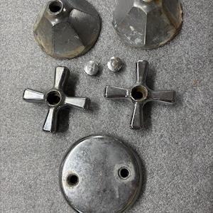 May include: A collection of vintage bathroom hardware components, including two faceted escutcheons, two cross-shaped faucet handles, two small screws, and a round drain cover. The pieces are made of metal and show signs of wear.