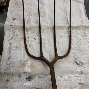 May include: An antique, rusty metal pitchfork with four tines. The pitchfork is lying on a white, patterned cloth. The tines are long and slender, and the handle is a single piece of metal. The overall appearance suggests a tool used for agricultural purposes.