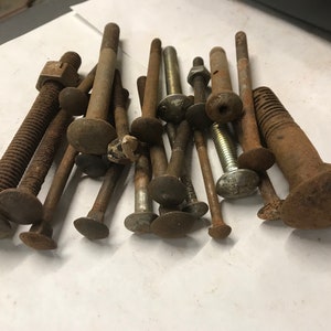 May include: A collection of various rusty bolts and screws with different head shapes and sizes. Some have nuts attached. The metal fasteners are arranged on a white surface, showing signs of age and wear.