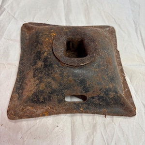 May include: A square, rusty metal object with a central circular opening and a rectangular hole on one side. The object has a dark, weathered appearance with patches of orange rust.