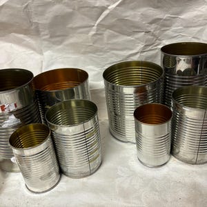 May include: A set of seven silver metal cans of varying sizes. The cans are all empty and have a shiny, reflective surface.