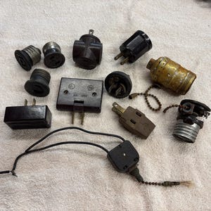 May include: A collection of vintage electrical components, including plugs, sockets, and a lamp holder. The items are in various shades of black, brown, and gold, with signs of wear.