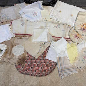 May include: A collection of vintage linens, including embroidered doilies, napkins, and placemats. The textiles feature floral and geometric patterns in white, cream, red, and yellow. Some pieces have lace trim.