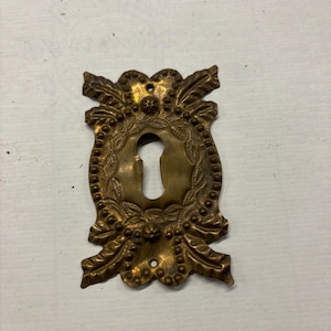 May include: A vintage brass keyhole cover with an ornate design featuring leaves and beads. The keyhole is oval shaped and the cover has a worn, antique finish.