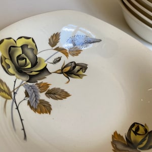 May include: A white ceramic bowl with a floral design featuring yellow and brown roses. The bowl has a slightly squared shape and a smooth, glossy finish.