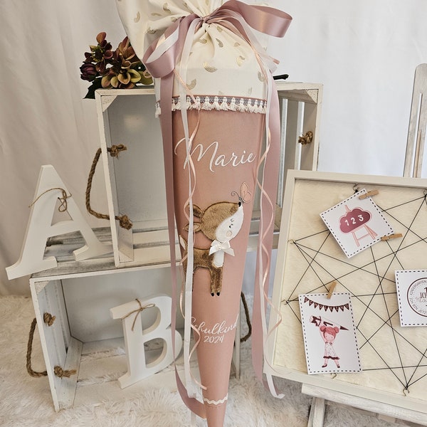 Può includere: Un cono scolastico rosa e bianco con un'applicazione a forma di cervo e il nome "Marie" in corsivo. Il cono è decorato con una parte superiore in tessuto bianco con dettagli dorati e un nastro rosa. È visibile anche il testo "Schulkind 2024".